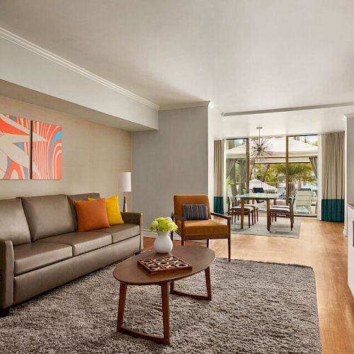 A modern living room with a sofa, armchairs, coffee table, flat-screen TV, and a vibrant wall art; leads to a dining area with outdoor views.