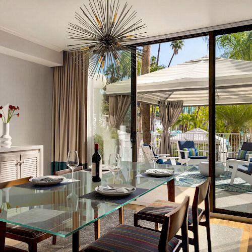 A dining area with a glass table set for two, a wine bottle, a large window, and an outdoor seating area with a canopy and view of palm trees.