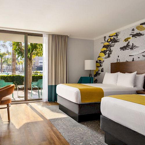 A hotel room with two beds, yellow accents, a mural, a TV, a desk, a chair, and a view through glass doors of greenery outside.