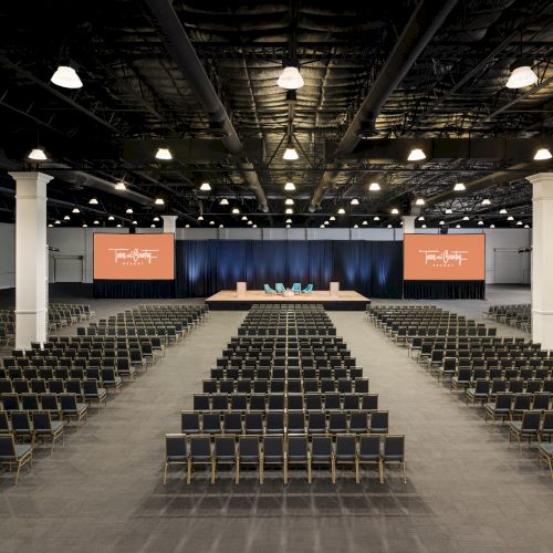 The image shows a large, empty conference hall with rows of chairs facing a stage and screens on the sides for presentations.