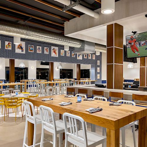 The image shows an empty sports bar with wooden tables, white and yellow chairs, sports decor, and a TV displaying a football game.