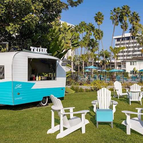 A turquoise and white camper is parked on a lawn surrounded by white Adirondack chairs; there are palm trees and buildings in the background.