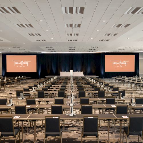 The image shows a large conference room with rows of tables and chairs facing a stage with two screens displaying similar messages.
