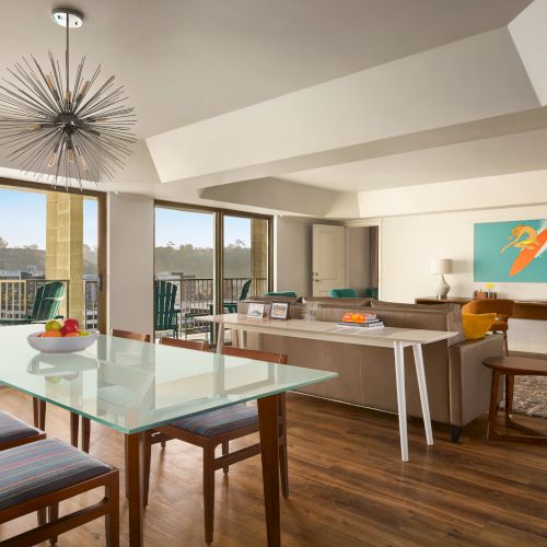 A modern open-plan living room features a dining table, stylish chairs, a sofa, contemporary art, and large windows with natural light.