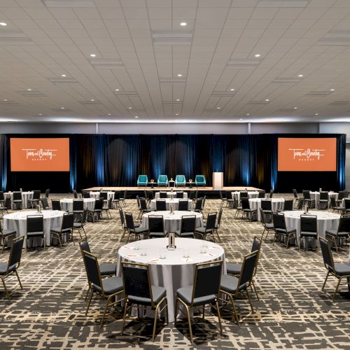 A large conference room with round tables, chairs, and two screens flanking a stage with five chairs and a podium, set for an event.