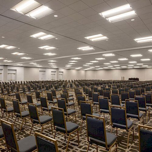 An empty conference hall with rows of chairs neatly arranged facing a stage area under bright overhead lights.