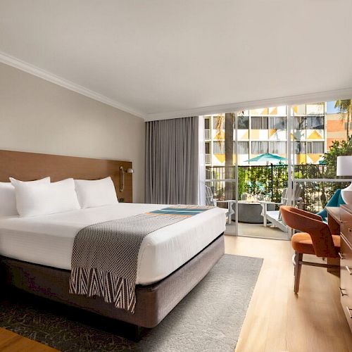 This is a photo of a modern hotel room with a large bed, a TV, a desk and chair, a balcony with a view of palm trees and outdoor seating.