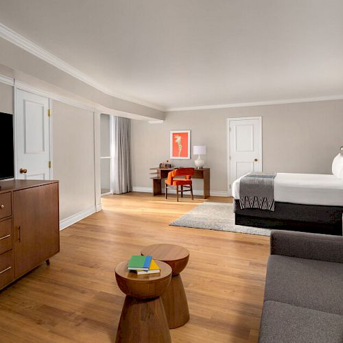 A spacious modern hotel room featuring a large bed, sofa, TV, desk, and wooden furniture, all set on a hardwood floor.