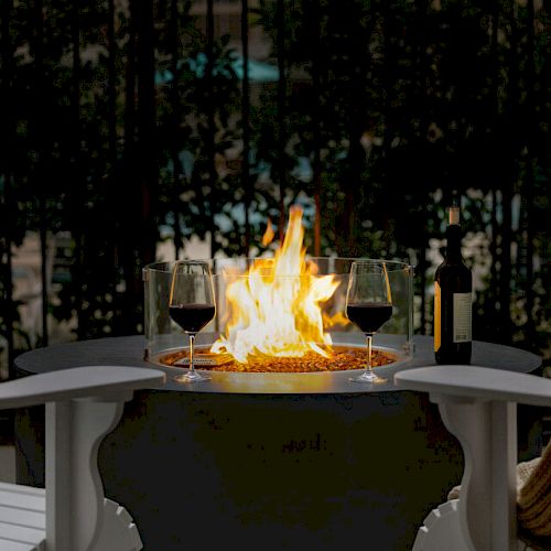A cozy setup with a fire pit, two glasses of red wine, a bottle of wine, and chairs, one draped with a blanket, in an outdoor setting.