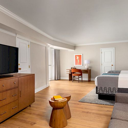 A spacious hotel room features a large TV, a wooden dresser, two beds, a sofa, a small round table, and a desk with a chair.