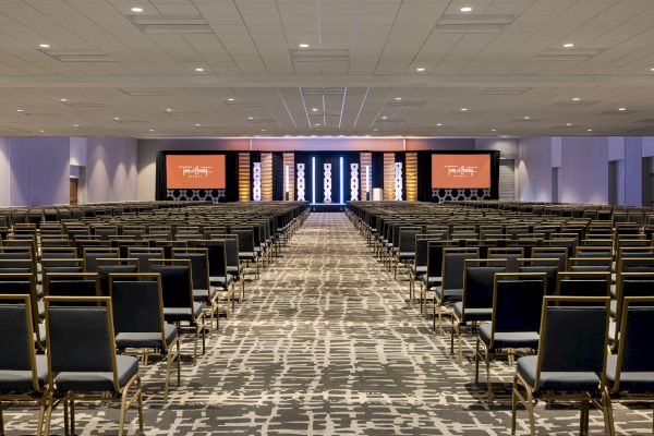 A spacious conference room with numerous rows of empty chairs facing a stage with large screens and modern lighting. Convention center-style hotel in Mission Valley.