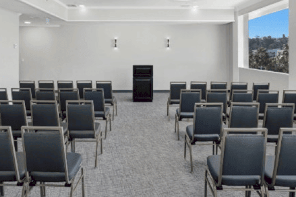 A small conference room with rows of gray chairs facing a podium at the front, illuminated by ceiling lights and natural light from a large window.