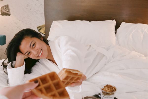A person is relaxing in bed, smiling, while another hand offers waffles. A tray with breakfast items is also on the bed.