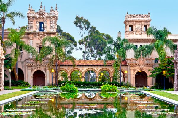 This image depicts an ornate, historic building with Mediterranean-style architecture, surrounded by palm trees and reflected in a tranquil pond.