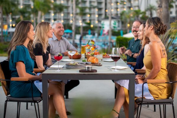 A group of six people are sitting at an outdoor dining table, enjoying food and drinks, with city lights and trees in the background.