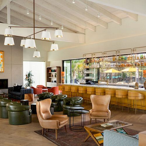 A modern lounge bar with leather seating, a fireplace, art on the wall, and a well-stocked bar area under high ceilings and stylish lighting.
