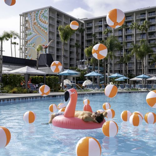 A person lounges on a pink flamingo float in a pool surrounded by numerous orange and white beach balls.
