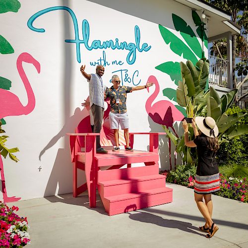 Two people stand on a pink platform posing for a photo; another person takes their picture in front of a mural with flamingos and the words "Flamingle with me."