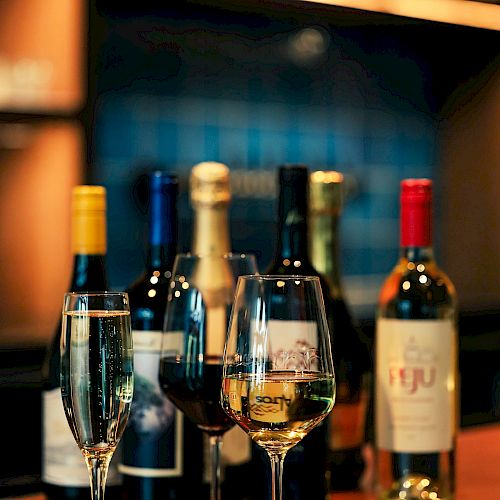 Various bottles of wine and filled wine glasses are arranged on a wooden bar counter with a blurred background.