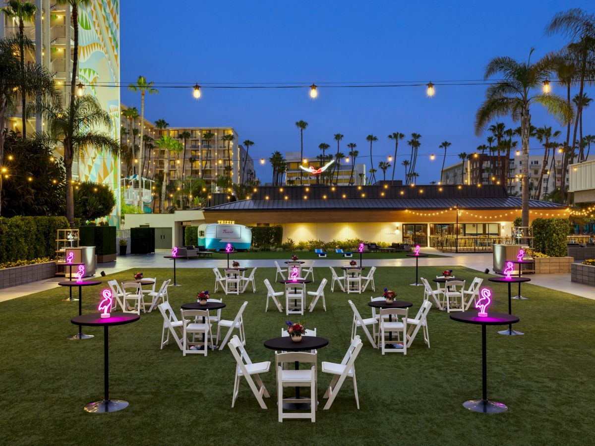 An outdoor event space is set up with round tables and chairs on a lawn, surrounded by string lights and palm trees, at dusk. Ballrooms, breakout rooms, and outdoor venues in San Diego.