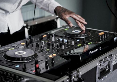A DJ is mixing music on a Pioneer DJ controller with various knobs and buttons being adjusted. The DJ's hand is on one of the jog wheels.