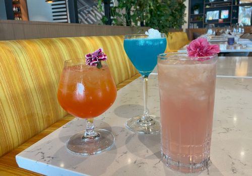 The image shows three colorful cocktails on a table, each garnished with a flower, in a bright and modern restaurant setting.