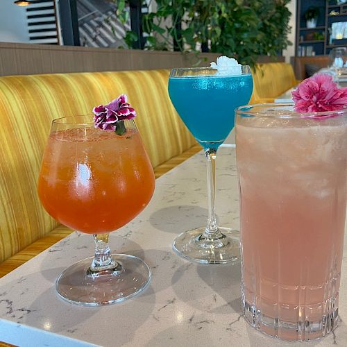 The image shows three colorful cocktails on a table, each garnished with a flower, in a bright and modern restaurant setting.