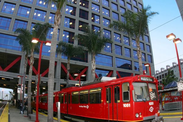 A red trolley is at a station with an urban building and palm trees in the background.