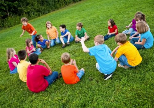 A group of children wearing colorful shirts sit in a circle on a grassy field, engaging in outdoor activities together.