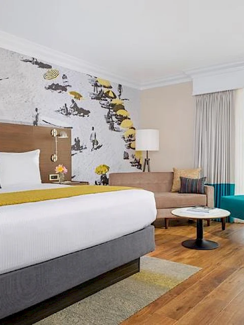 A modern hotel room with a large bed, colorful seating, and wall art, features a sliding glass door leading to a balcony with a greenery view.