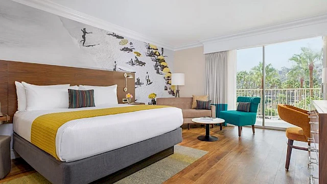 A modern hotel room with a large bed, colorful seating, and wall art, features a sliding glass door leading to a balcony with a greenery view.