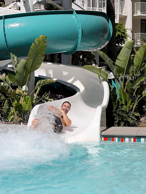 A person sliding down a water slide into a pool, surrounded by plants and palm trees, with water splashing around them.