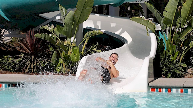 A person sliding down a water slide into a pool, surrounded by plants and palm trees, with water splashing around them.