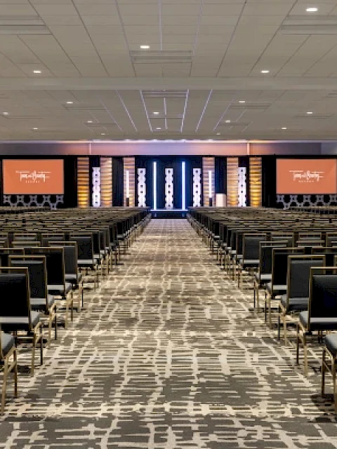 An empty conference room with rows of chairs facing a stage, featuring two screens displaying a logo.