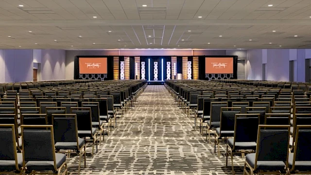 An empty conference room with rows of chairs facing a stage, featuring two screens displaying a logo.