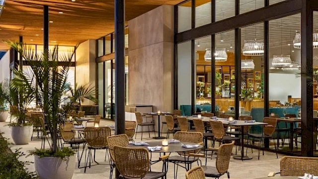 An outdoor dining area with empty tables and chairs, potted plants, and large windows reflecting the interior lights of a building.