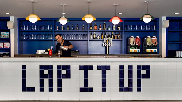 A bartender is serving drinks at a bar with "LAP IT UP" in large letters on the front.