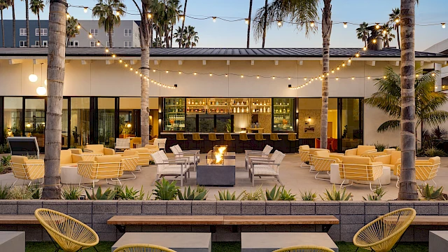 Outdoor patio with yellow chairs, fire pit, string lights, and palm trees, leading to a bar with a modern design at dusk.