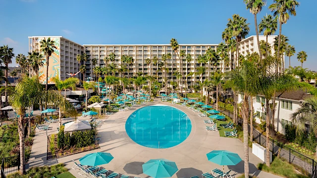 A hotel with a large circular pool, surrounded by blue sun loungers and umbrellas, set amidst palm trees under a clear blue sky.