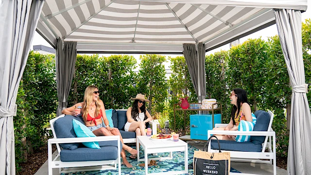 Three people relax under a striped canopy, sitting on blue chairs, with a table and a decorative rug beneath them.