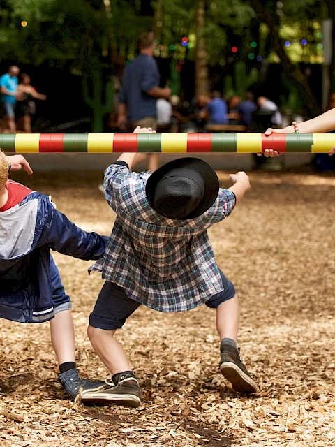 Two children participate in a limbo game, bending backwards under a colorful pole held by a woman in a festive setting.