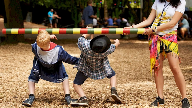 Two children participate in a limbo game, bending backwards under a colorful pole held by a woman in a festive setting.