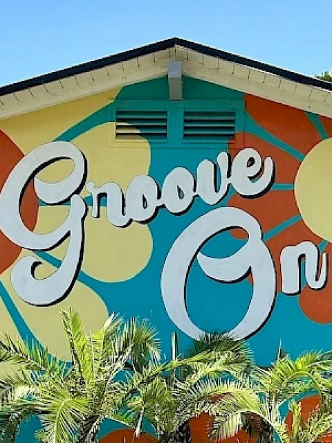 A colorful mural with the words "Groove On" and retro-style floral patterns decorates a building, surrounded by palm trees.