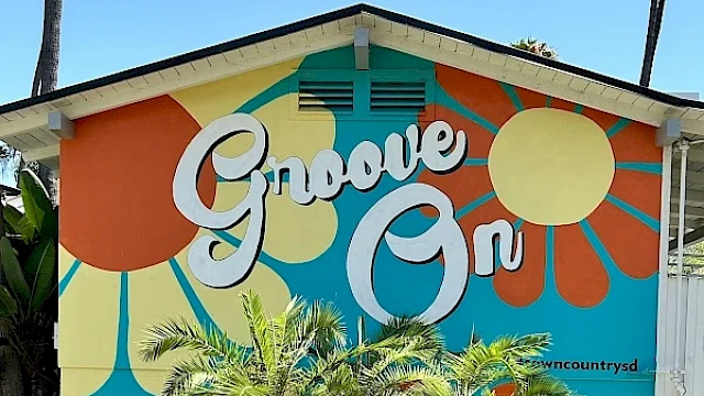 A colorful mural with the words "Groove On" and retro-style floral patterns decorates a building, surrounded by palm trees.